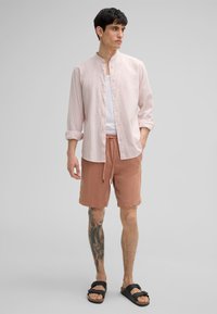 Camicia a righe rosa chiaro con bottoni, canotta bianca e pantaloni corti color ruggine con cordino. Sandali neri. Modella con un tatuaggio su una gamba.