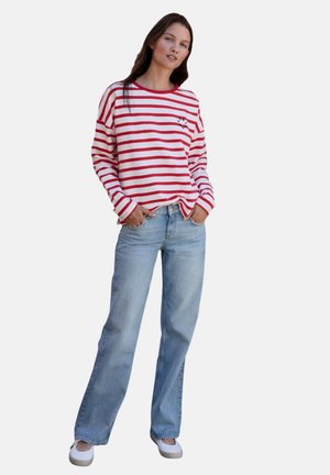 Frau steht mit den Händen in den Taschen, trägt ein rot-weiß gestreiftes Langarmshirt, hellblaue Jeans und weiße Schuhe vor einem schlichten Hintergrund.