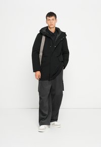 Zwarte waterdichte parka met een capuchon, voorzien van grijze accenten, twee voorzakken en een relaxte pasvorm. Gecombineerd met losse grijze broeken en sneakers.