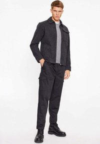 Giacca grigia effetto tessuto con colletto, abbinata a pantaloni cargo neri. Stivaletti neri con lacci completano l'outfit, enfatizzando uno stile utilitaristico.