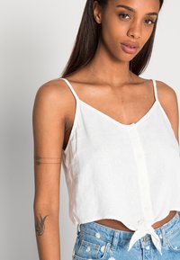 Top de linho branco, curto, com alças finas, decote em V, fecho frontal com botões e detalhe de ponta de nó na bainha. Tecido leve e respirável.