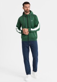 Mann in grüner Zip-Hoodie mit weißen Streifen an den Ärmeln, blauen Jeans und weißen Sneakern, der vor einem weißen Hintergrund steht.