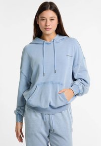 Gaiši zils kapuces džemperis izgatavots no mīksta auduma, ar kanguru kabatu, pievilktņu kapuci un ribotām aprocēm. Tam ir neliela logo uz krūtīm.