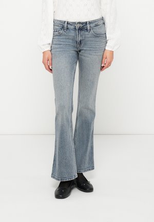 Svetlo modri flared jeans z srednjim pasom, subtilnim zbledeljem in kontrastnim šivanjem. Nosi jih z črnimi balerinkami.