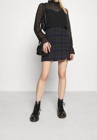 Svart genomskinlig långärmad topp med ett texturerat mönster, tillsammans med en marinblå rutmönstrad minikjol med gröna och röda linjer. Svarta snörkängor.