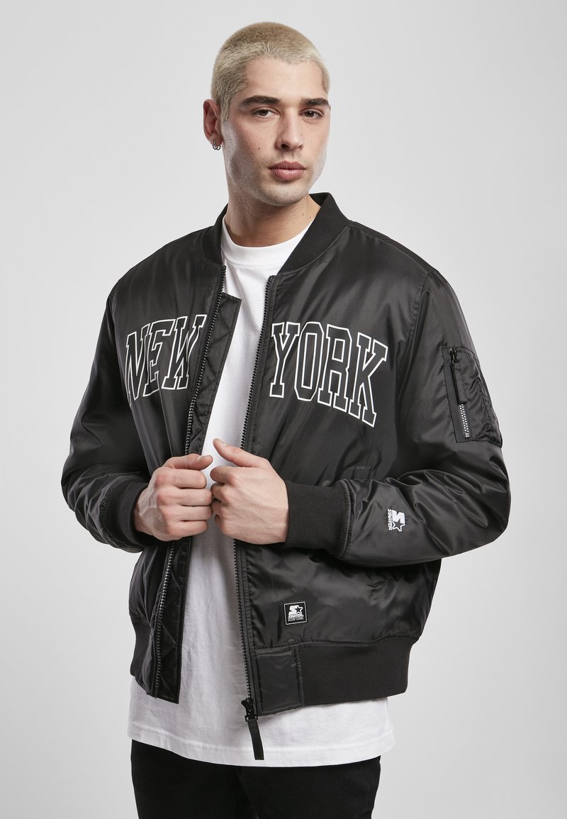 Chaqueta bomber negra con letras "NEW YORK". Fabricada en tejido brillante, presenta un bolsillo con cremallera, puños acanalados y un parche con el logo.