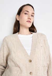 Beige kötött kardigán V-kivágással, két elülső zsebből és barna gombos záródással, egy fehér ing felett.
