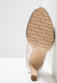 Chaussure à talon aigu en rose clair avec une finition satinée lisse, présentant une pointe en amande et une semelle texturée portant l'inscription "Tamaris" dans un motif répétitif.