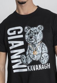 Crna majica s velikim grafičkim printom medvjeda ispunjenog dizajnom novčanica. Ispod se u podebljanim bijelim slovima ispisuje "GIANNI KAVANAGH".
