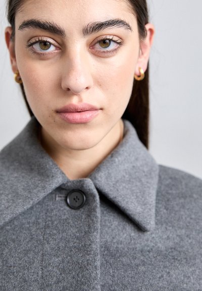 Gray wool coat with a wide collar, single black button closure, and smooth texture; model has light skin and gold hoop earrings.