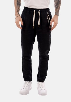 Uomo che indossa pantaloni jogger neri con coulisse bianche, un accessorio a catena, scarpe da ginnastica bianche e braccia tatuate su sfondo neutro.