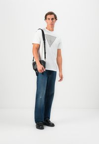T-shirt blanche avec un logo graphique gris, jeans droit bleu et chaussures en cuir noires. Sac bandoulière noir avec un détail de texte en relief.