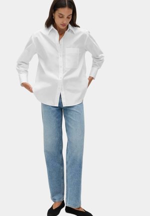 Femme portant une chemise blanche ample à boutons, un jean droit bleu clair et des chaussures noires sans lacets, debout les mains dans les poches.