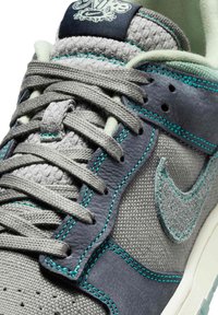 Lähikuva harmaasta Nike-tennarista, jossa on kudottu kangas, mokkanahkaiset päällysteet, turkoosit tikit ja nyörit solmittuna pehmustetun kielen yli, jossa on Nike-logo.