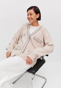 Cardigan beige con ribete azul marino, ajuste oversized, dos bolsillos frontales y cierre de botones. Modelo sentado en una silla negra, vistiendo pantalones blancos.