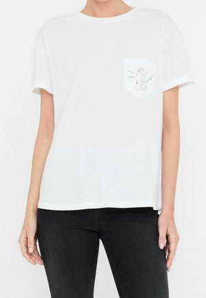 Persona que lleva una camiseta blanca lisa de manga corta con un pequeño diseño decorativo en el bolsillo del pecho izquierdo y pantalones negros.