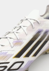 White soccer cleat with black and gold stripes, featuring a textured upper and lace closure, accented by purple and gold highlights.