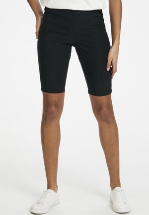 Person trägt schwarze, knieumspielende, eng anliegende Shorts, weiße Turnschuhe und ein weißes Oberteil und steht mit einer Hand in der Hüfte vor einem weißen Hintergrund.