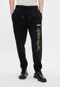 Μαύρο φόρεμα με ελαστική μέση, με μπλε λογότυπο και κίτρινο κείμενο "Jujutsu Kaisen" κατά μήκος του αριστερού ποδιού. Λείος υφασμάτινος χαρακτήρας.