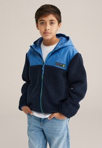 Polárdzseki kék steppelt felsőrésszel, sötétkék testtel, zöld cipzárral és két elülső zsebbel. Fehér póló és világos farmer fölött viselve.