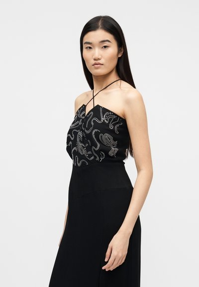 Νεαρή γυναίκα με μακριά σκούρα μαλλιά, ντυμένη με μαύρο φόρεμα τύπου halter με ασημένια σχέδια από χάντρες στο κορσάζ, στέκεται μπροστά σε απλό φόντο.