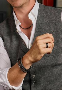 Grijze textuurvest over een wit shirt, geaccentueerd met een zwarte kralenketting, drie zwarte leren armbanden en een zwarte ring.
