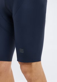 Ναυτικά μπλε ποδηλατικά σορτς με διακριτική ραφή και μικρό ανακλαστικό λογότυπο πάνω από το γόνατο στο πόδι ενός ατόμου.