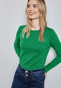 Långärmad grön topp, gjord av slät tyg, kombinerad med en mörkblå denimkjol med knappdetaljer och kontrasterande söm.