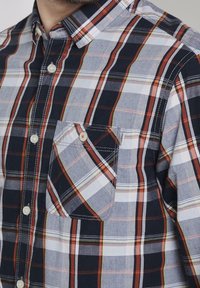 Chemise à carreaux avec des teintes bleu marine, gris et orange. Fabriquée en tissu léger, avec une poche poitrine et des boutons blancs.