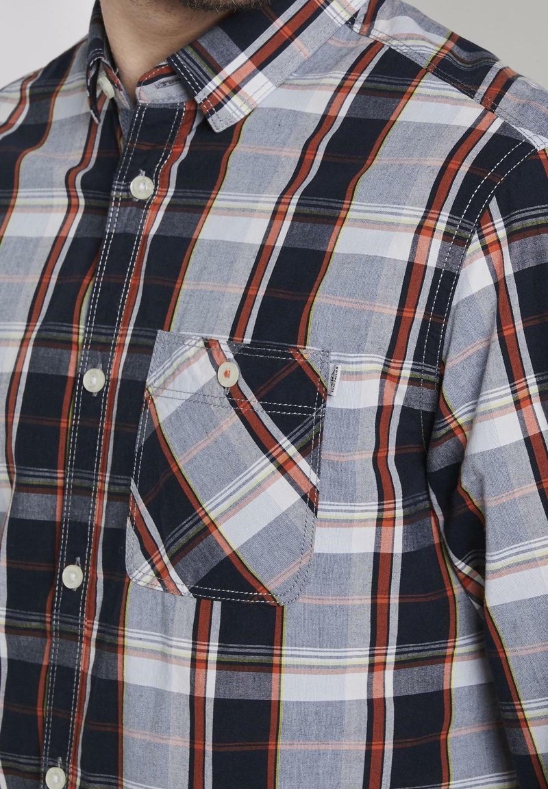 Chemise à carreaux avec des teintes bleu marine, gris et orange. Fabriquée en tissu léger, avec une poche poitrine et des boutons blancs.