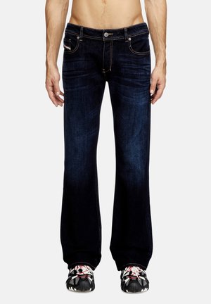 Homme portant un jean bootcut bleu foncé et des baskets noires et blanches, debout devant un fond clair uni.