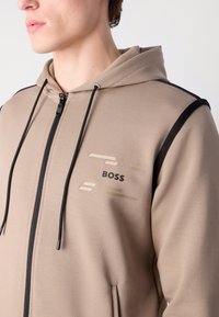 Sweatshirt zippé beige avec des accents et un logo noirs. Il dispose d'une capuche, de poches avant et d'un tissu texturé, présentant un design minimaliste.