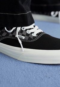 Svart lær-lav topp sneakers med et grått rutemønster, hvite snører og en gummisåle, plassert på en teksturert blå overflate.