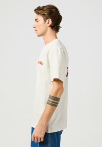 Camiseta de algodón blanca de manga corta, con texto rojo y negro en el frente. Combinada con jeans de mezclilla azul. Se muestra vista lateral.