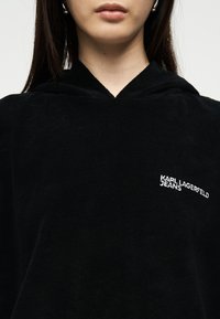 Γυναίκα που φοράει μαύρη μπλούζα με κουκούλα, με το κείμενο "KARL LAGERFELD JEANS" κεντημένο με άσπρα γράμματα στο στήθος, εμφανιζόμενη από τους ώμους έως τα χείλη.