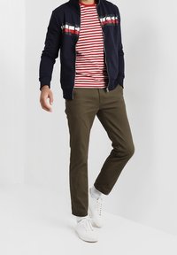Giacca navy con cerniera e dettaglio a righe, t-shirt a righe rosse e bianche, pantaloni verde oliva e sneakers bianche con calze a coste.