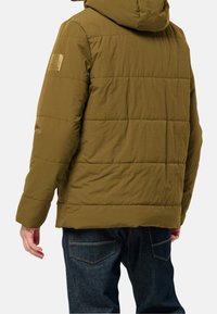 Olivgrön vadderad jacka med huva, med ett quiltat mönster och en logotyp på ärmen. Bärs med mörkblå denimjeans.