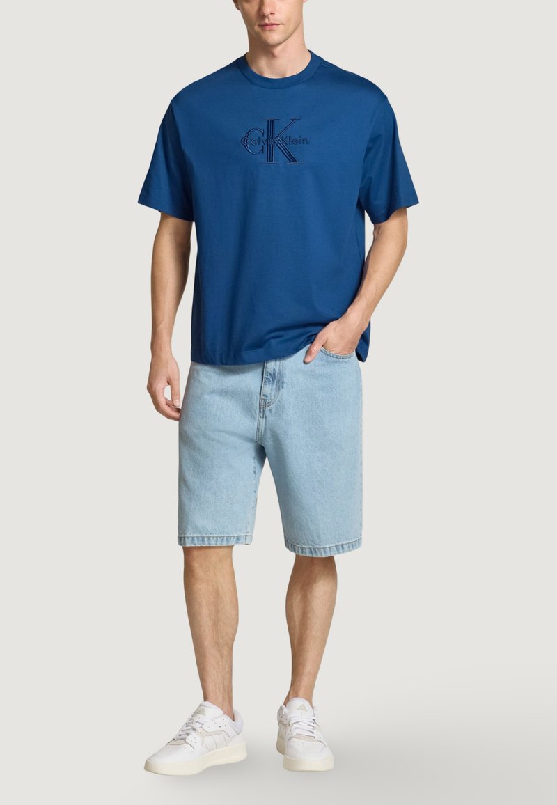 Modrá bavlněná tričko s logem, spárované se světle modrými džínovými šortkami. Model nosí bílé tenisky, což představuje ležérní outfit.