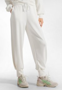 Weiße, locker sitzende Joggerhosen mit elastischen Bündchen und einem Kordelzugbund, kombiniert mit beigen Sneakers mit grünen Akzenten.