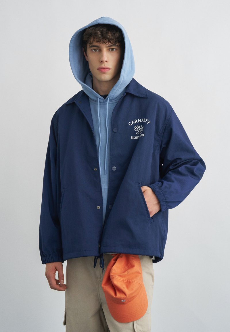 Giovane uomo con felpa con cappuccio azzurro sotto una giacca Carhartt blu navy, che tiene un cappello arancione, con pantaloni beige su uno sfondo semplice.