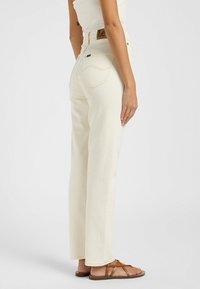 Jean en denim blanc avec une coupe droite, cinq poches et des détails de couture légers. Portés avec des sandales à lanières brunes.