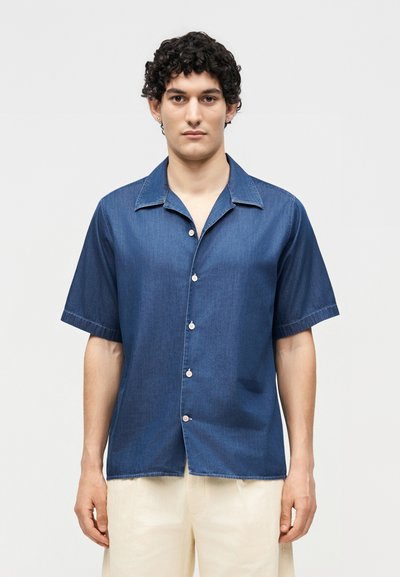 Jeune homme aux cheveux courts et bouclés portant une chemise bleu foncé à manches courtes boutonnée et un pantalon beige clair, debout devant un fond uni.