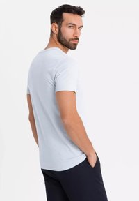 Vaaleansininen lyhythihainen t-paita, jossa on pyöreä pääntie ja sileä rakenne. Malli seisoo sivuttain käsi taskussa, esitellen rentoa istuvuutta.