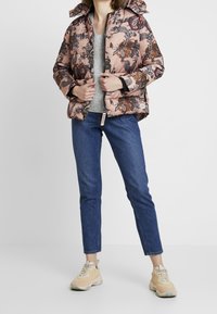 Chaqueta acolchada rosa con estampado floral, cierre de cremallera y capucha; combinada con un suéter gris, jeans azules y zapatillas color beige claro.