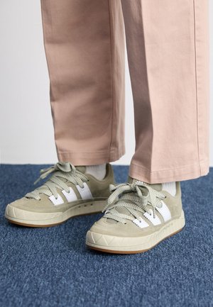 Voeten in lichtgroene en witte sneakers met dikke veters en beige broek staan op een blauw tapijt.