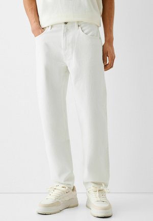 Personne portant un jean blanc ample avec les mains dans les poches et des baskets écrues, debout devant un fond blanc.