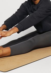 Persona estirada hacia adelante sentada en una esterilla de yoga, vistiendo una camiseta de manga larga oscura y leggings grises, alcanzando los pies con los brazos extendidos.