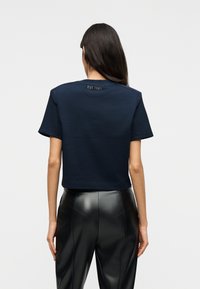 Γυναίκα με μακριά σκούρα μαλλιά που φοράει ένα μπλε ναυτικό κοντό μπλουζάκι με την επιγραφή "EST. 1981" στο πίσω μέρος του λαιμού και μαύρο δερμάτινο παντελόνι, στέκεται γυρισμένη από την πλάτη.