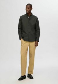 Camisa de botones verde oscuro con un bolsillo en el pecho, combinada con pantalones beige claros y zapatos negros. Tejido suave, corte ajustado, mangas largas.