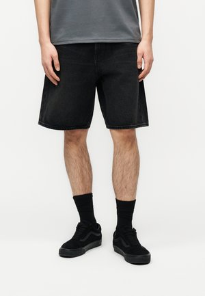 Uomo che indossa pantaloncini di jeans neri, calzini neri e scarpe da ginnastica nere, in piedi contro uno sfondo bianco semplice.